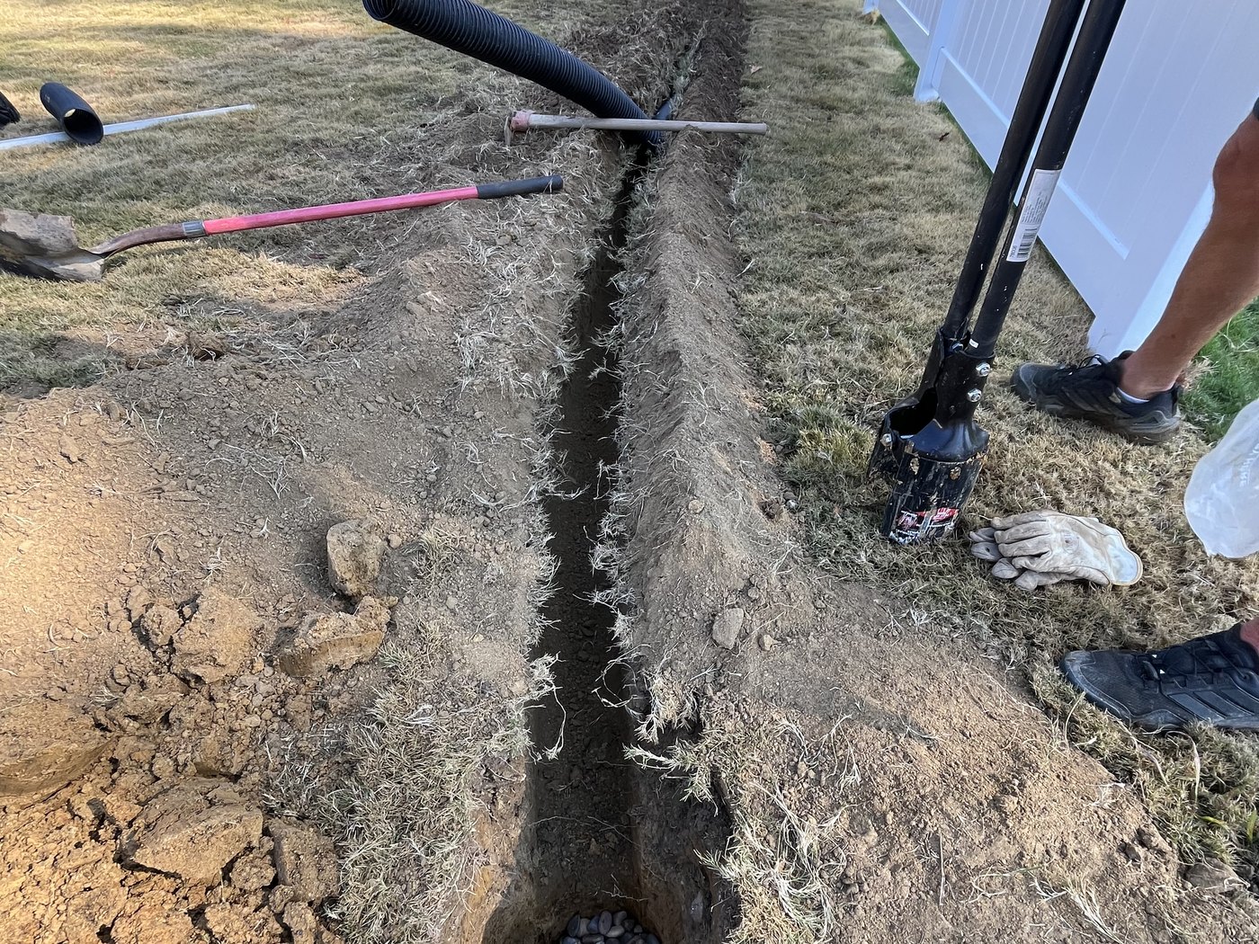 An open drainage trench with perforated pipe and a pocket of river rock alongside a fence line