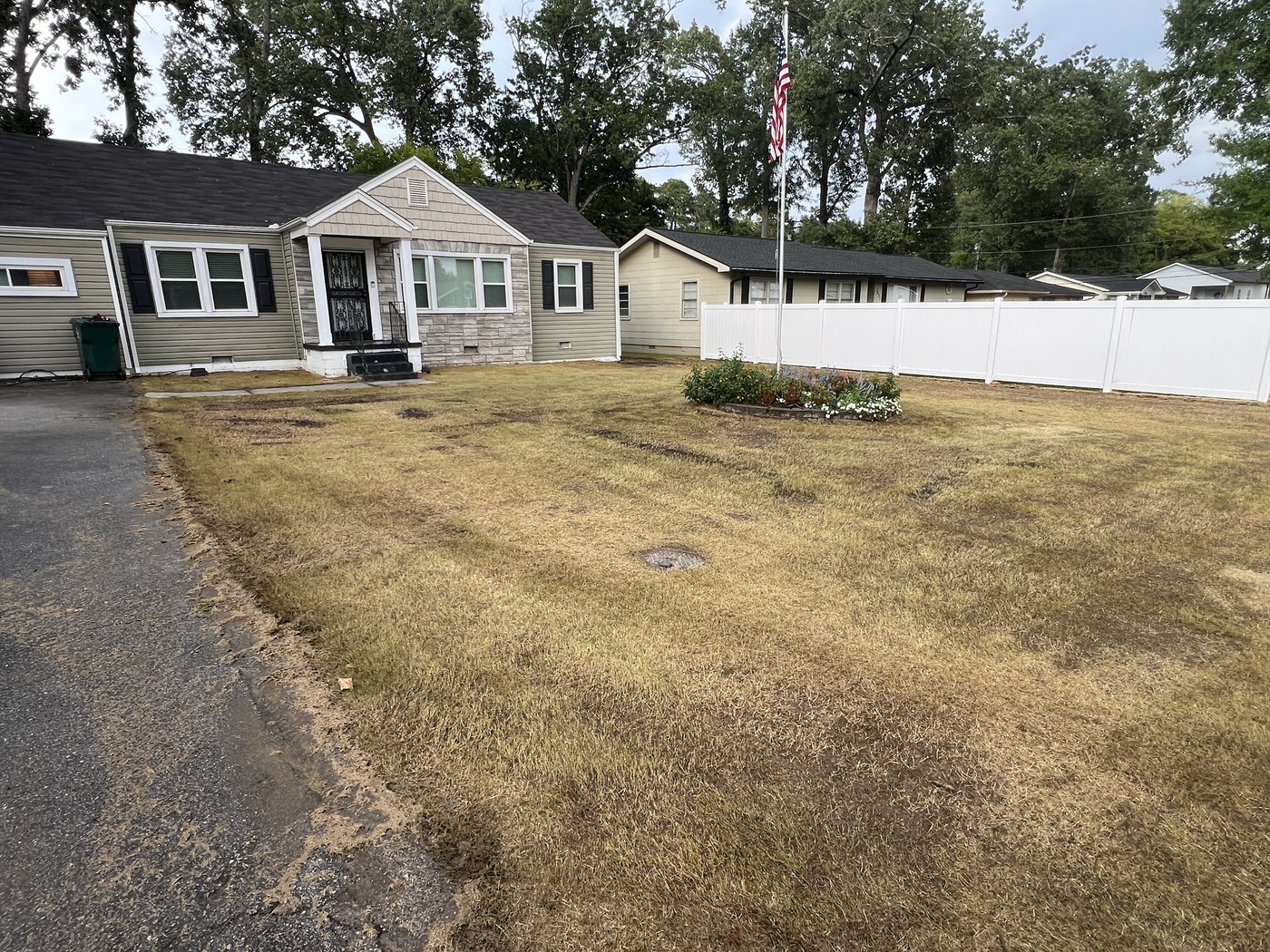 A front lawn of thin brown grass with bare patches showing soil, house and flagpole behind