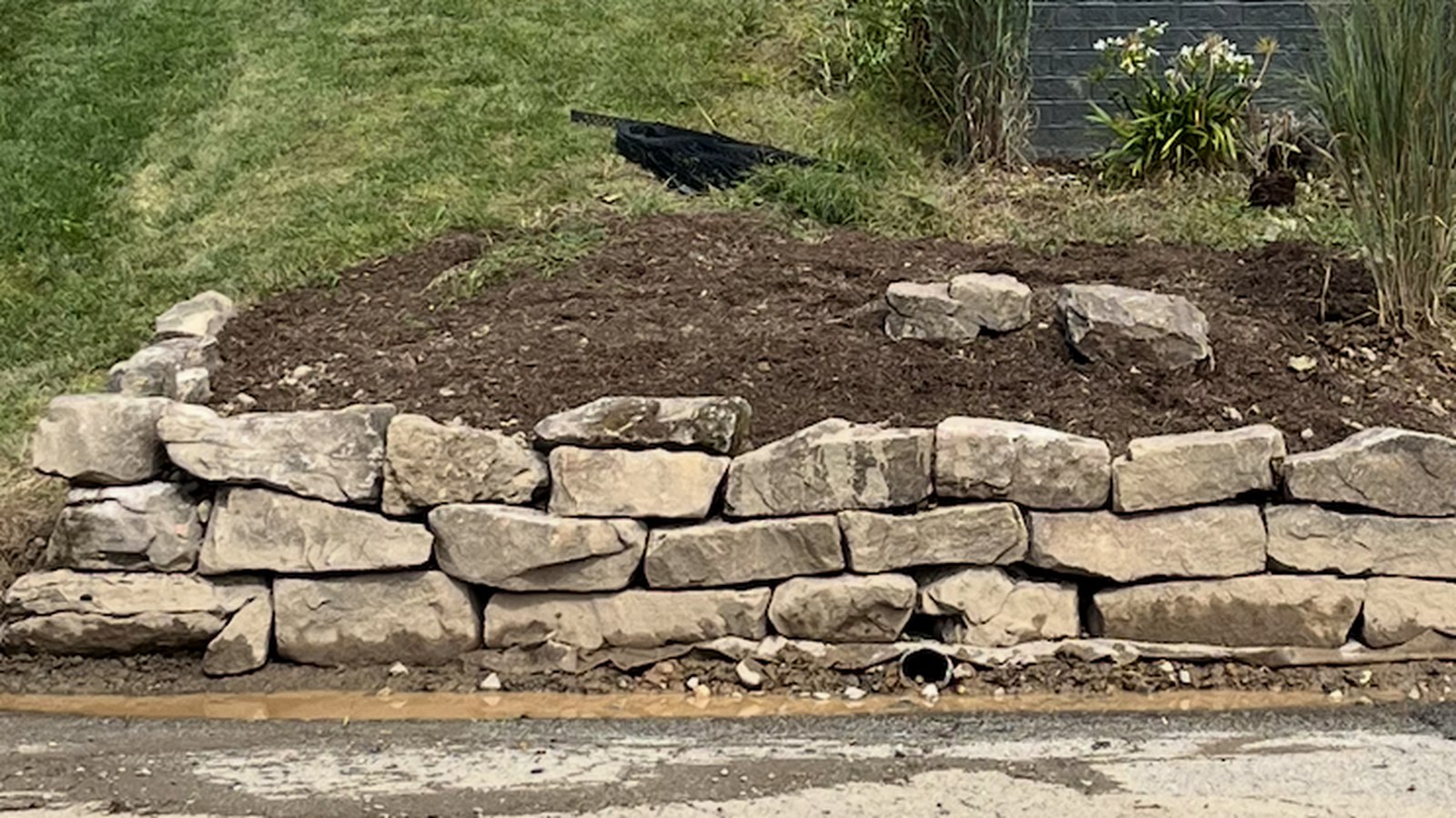 A dry-stacked limestone wall edging a raised mulched flower bed, with a striped lawn behind it