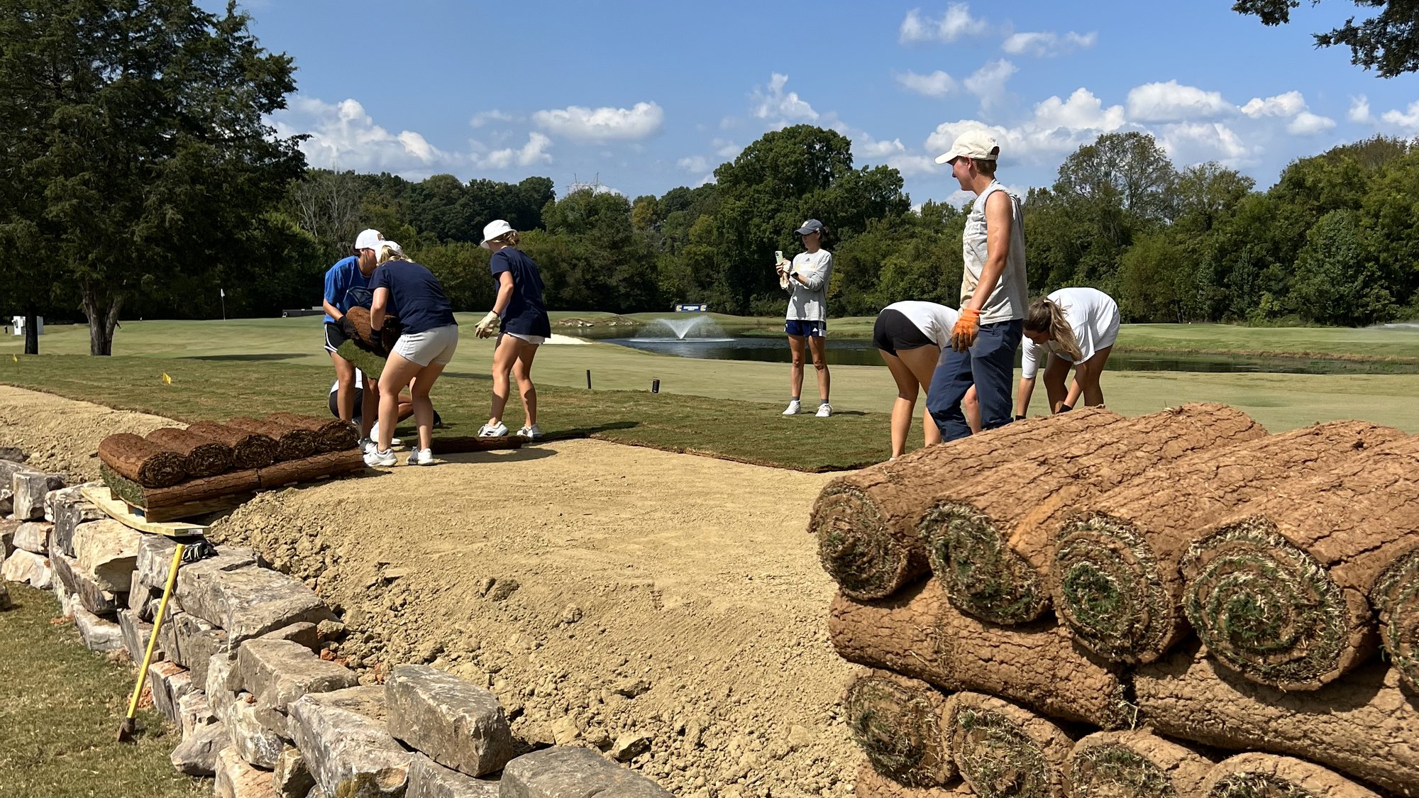 Pallets of fresh sod rolls stacked along a hand-laid stone wall on a golf course, with a crew working in the background