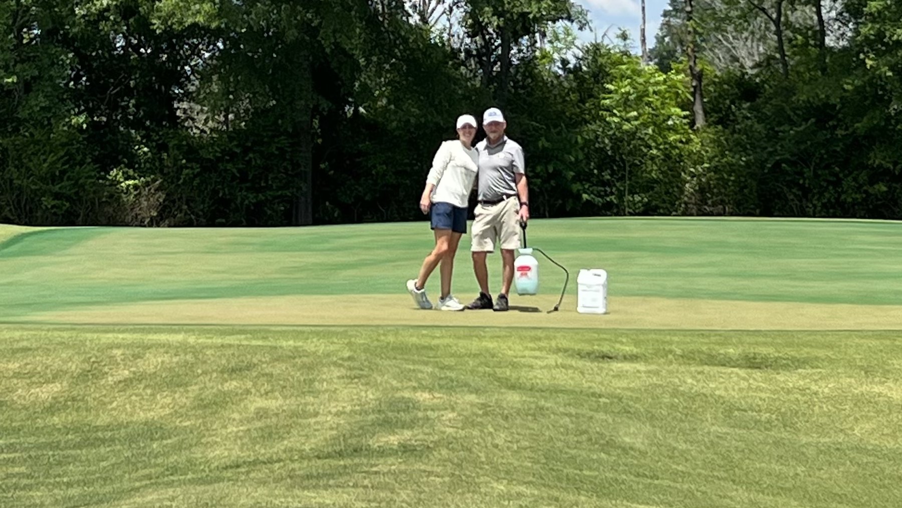 Chip and Colette on a putting green with a sprayer and a jug of product